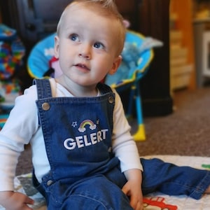 May include: A young child wearing a white long-sleeved shirt and blue denim dungarees with a rainbow and the word "GELERT" embroidered on the front. The child is sitting on a patterned rug and looking up.