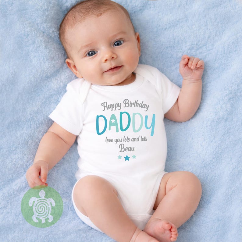 May include: A baby wearing a white onesie with the text "Happy Birthday DADDY" in gray and teal. The text "love you lots and lots Beau" is below the word "DADDY", with two blue stars. The baby is lying on a light blue blanket.