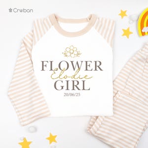 May include: A white long-sleeved top and matching trousers set with a beige and white striped pattern. The top features the text "FLOWER Elodie GIRL" with a flower graphic and the date 20/06/25.