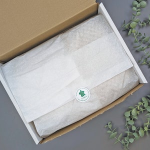 Puede incluir: Una caja de cart&oacute;n abierta con papel de seda blanco en el interior y una pegatina verde y blanca. La caja est&aacute; sobre una superficie gris con follaje verde en el fondo.