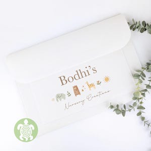 May include: A white nursery creation with a flap closure. The front displays the text "Bodhi's Nursery Creations" with illustrations of an elephant, bear, giraffe, sun, and flowers. A green turtle logo is in the lower left corner.