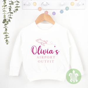 May include: White long-sleeved sweatshirt with a pink airplane graphic and the text "Olivia's Airport Outfit" in pink.