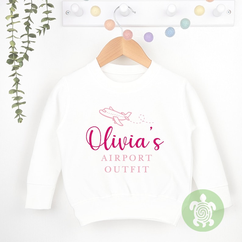May include: White long-sleeved sweatshirt with a pink airplane graphic and the text "Olivia's Airport Outfit" in pink.