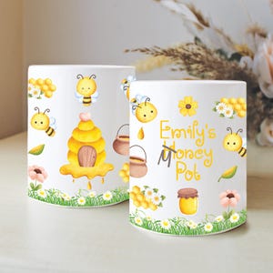 May include: Two white ceramic mugs with a bee-themed design. One mug features a beehive, bees, and flowers. The other mug has the text "Emily's Money Pot" with bees, flowers, and a honey jar. The mugs are decorated with a cartoon style.