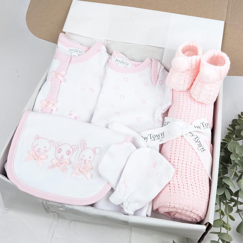 May include: A baby gift set in a box containing a white long-sleeve onesie with pink trim, a white short-sleeve onesie with a pink heart pattern, a white bib with embroidered animal faces and pink bows, white mittens, and a pink knit blanket with booties.
