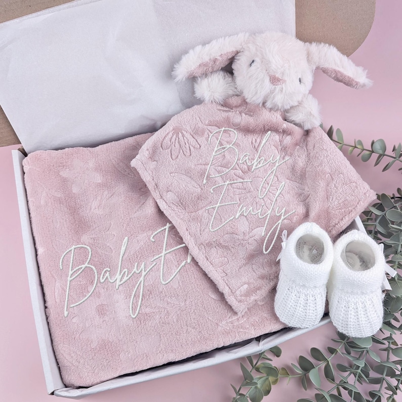 May include: A baby gift set in a white box, featuring a pink blanket and a matching security blanket with floral embroidery and the name "Baby Emily". Includes a white plush bunny and a pair of white baby booties.
