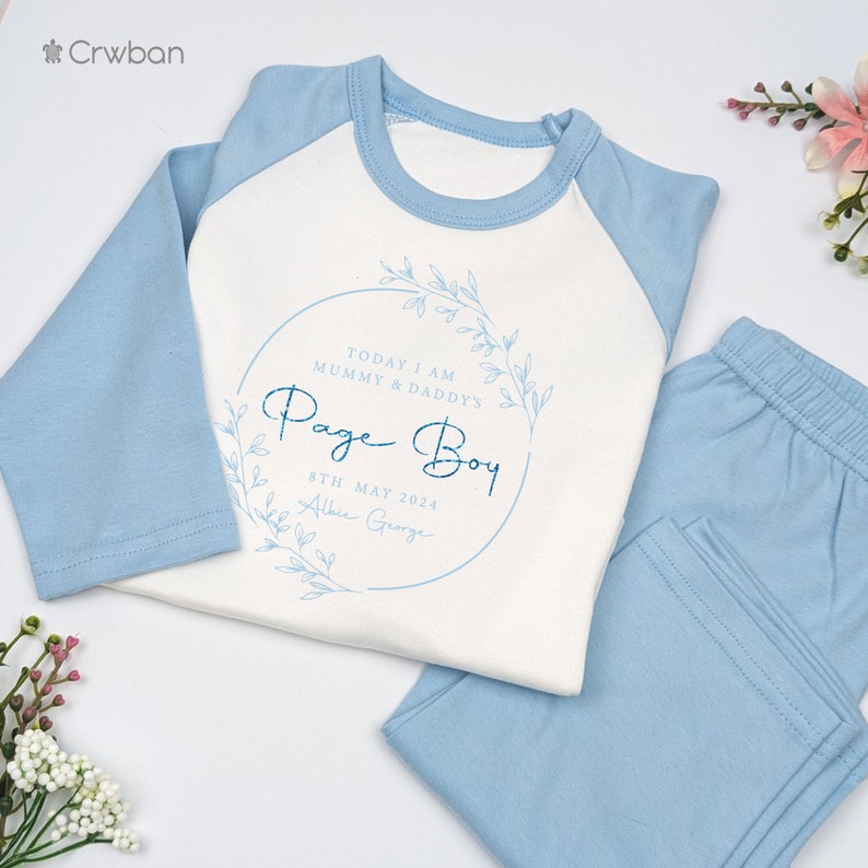 May include: A two-piece baby outfit in light blue and white. The long-sleeved top has blue sleeves and a white body with the text "Page Boy" inside a floral wreath. The matching pants are light blue. The text also includes the date "8th May 2024".