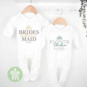 May include: Two white baby sleepsuits suspended on wooden hangers. One reads "BRIDES Tallulah MAID" with a floral design and the date 12/09/25. The other reads "FLOWER Elodie GIRL" with a floral design and the date 20/06/25.
