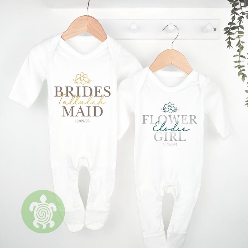 May include: Two white baby sleepsuits suspended on wooden hangers. One reads "BRIDES Tallulah MAID" with a floral design and the date 12/09/25. The other reads "FLOWER Elodie GIRL" with a floral design and the date 20/06/25.
