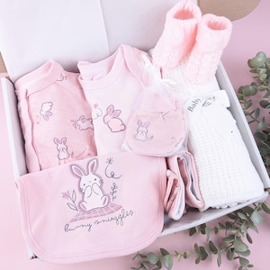 May include: A baby gift set in a white box containing a pink long-sleeve onesie, a pink bodysuit with a bunny, pink knit booties, a white textured blanket with a Baby tag, and a pink bib with a bunny graphic and the text bunny snuggles.