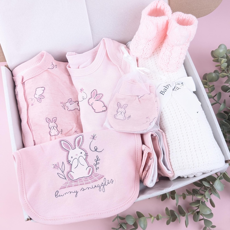May include: A baby gift set in a white box containing a pink long-sleeve onesie, a pink bodysuit with a bunny, pink knit booties, a white textured blanket with a Baby tag, and a pink bib with a bunny graphic and the text bunny snuggles.