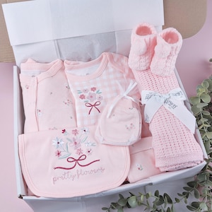 May include: A baby gift set in a white box containing a light pink floral onesie, a pink gingham bodysuit with a floral bouquet graphic, a pink bib embroidered with flowers and the text pretty flowers, pink knit booties, and a pink blanket.