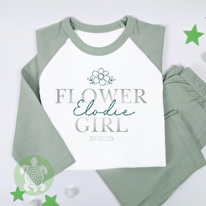 May include: A two-piece pyjama set in white and sage green. The top has a white front with sage green sleeves and collar. The text "FLOWER GIRL Elodie" is printed on the front, along with a flower graphic and the date 20/06/25.
