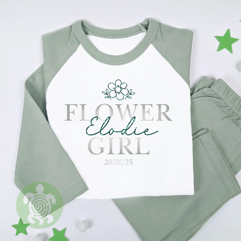May include: A two-piece pyjama set in white and sage green. The top has a white front with sage green sleeves and collar. The text "FLOWER GIRL Elodie" is printed on the front, along with a flower graphic and the date 20/06/25.
