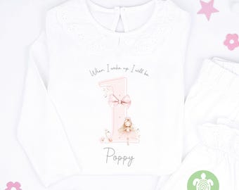 May include: White long-sleeved shirt with a lace collar and the text "When I wake up I will be" above a pink number one with a bow, a fairy, and the name "Poppy". Matching white pants are also visible.