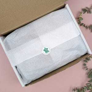 May include: Open cardboard box with white tissue paper and a green turtle sticker. The box is lined with white paper and contains a wrapped item. The background is a light pink surface with green leaves.