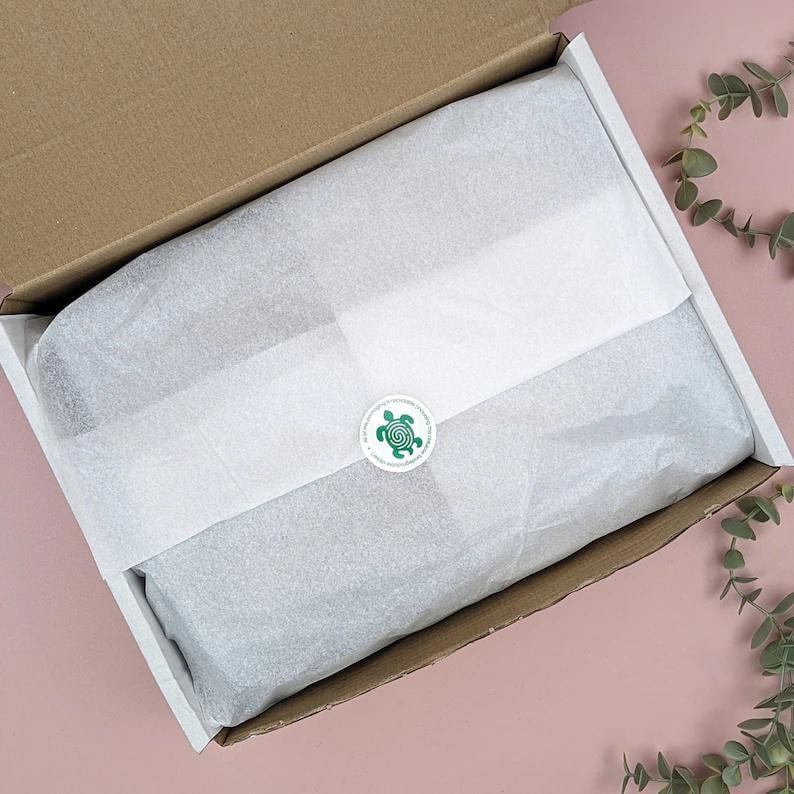 May include: Open cardboard box with white tissue paper and a green turtle sticker. The box is lined with white paper and contains a wrapped item. The background is a light pink surface with green leaves.