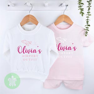 May include: Two white and pink t-shirts with a pink airplane graphic and the text "Olivia's Airport Outfit" on them. The shirts are hanging on wooden hangers.