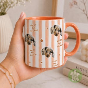 May include: A ceramic mug with a peach and white vertical striped pattern, featuring repeated images of a dapple dachshund puppy and the text World's Best Dog Mum interspersed with small star icons. The mug has a solid peach handle and interior.