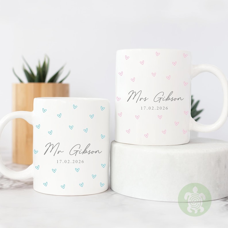 May include: Two white ceramic mugs with heart patterns. One mug has blue hearts and the text "Mr Gibson 17.02.2026". The other mug has pink hearts and the text "Mrs Gibson 17.02.2026". Both mugs have a curved handle.