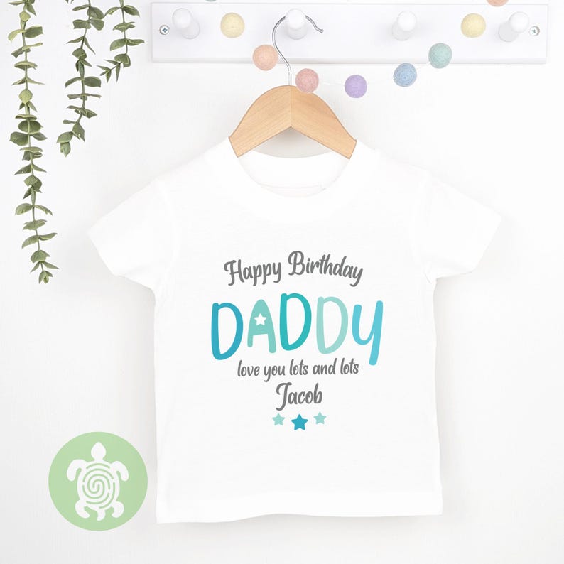 May include: White t-shirt with the text "Happy Birthday DADDY" in gray and blue. The shirt also includes the text "love you lots and lots Jacob" with blue star accents. The shirt is hanging on a wooden hanger.