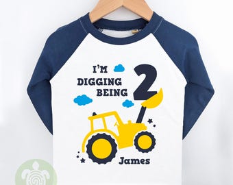 May include: White and navy blue long-sleeved shirt with a yellow construction vehicle graphic. The shirt features the text "I'm Digging Being 2" and a name below the graphic. A fun birthday shirt.
