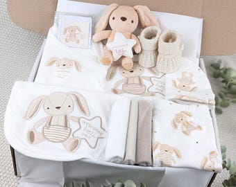 Può includere: Un set regalo per neonati in una scatola di cartone contenente un coniglio di peluche beige, un paio di scarpine in maglia color crema, una coperta bianca, un berretto bianco e indumenti bianchi, tutti con un disegno di coniglio e la scritta Welcome little one.
