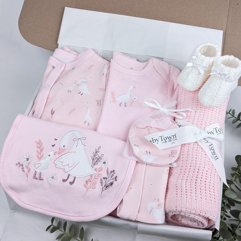 May include: A baby gift set in a box containing a pink long-sleeve onesie, a pink footed sleeper, a pink bib with an embroidered swan and gosling, a pair of white knit booties, and a pink cellular blanket tied with a BabyTown branded ribbon.
