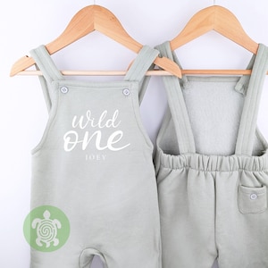 May include: A light green baby overall with the text "Wild One" and the name "Joey" printed on the front. The overall has adjustable straps and a pocket on the back.