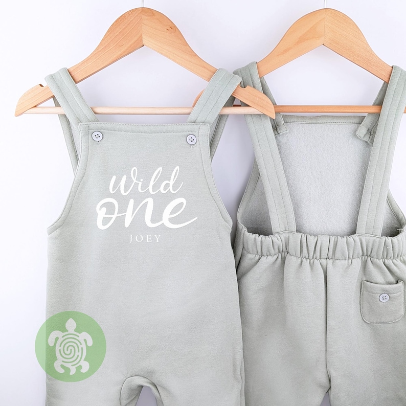 May include: A light green baby overall with the text "Wild One" and the name "Joey" printed on the front. The overall has adjustable straps and a pocket on the back.