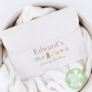 May include: A cream-coloured baby blanket with a decorative label featuring the text "Edward's Nursery Creations." The label includes illustrations of an elephant, bear, giraffe, sun, and flowers. The blanket is folded and placed in a woven basket.