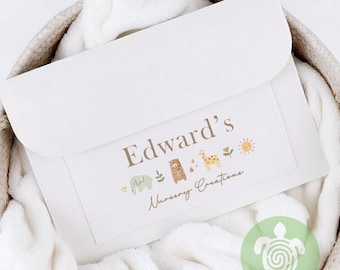 Puede incluir: Una manta de bebé de color crema con una etiqueta decorativa con el texto "Edward's Nursery Creations". La etiqueta incluye ilustraciones de un elefante, un oso, una jirafa, un sol y flores. La manta está doblada y colocada en una cesta tejida.