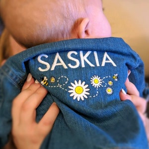 May include: A blue denim jacket with the name "Saskia" embroidered on the back. The embroidery is white and includes a line of small white daisies and yellow bees.