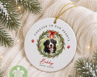 May include: A round white ceramic ornament featuring a photo of a Bernese Mountain Dog inside a green wreath with a red bow. The text reads FOREVER IN OUR HEARTS, Bobby, and 2015-2025, with red paw prints on the sides and a gold hanging string.