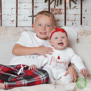 May include: A white short-sleeved t-shirt and white long-sleeved onesie featuring red and grey text. The onesie displays a red Christmas tree graphic with the text "Maeve's FIRST CHRISTMAS 2025". The t-shirt has partial text reading "CHRISTMAS".