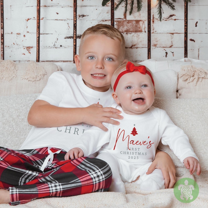 May include: A white short-sleeved t-shirt and white long-sleeved onesie featuring red and grey text. The onesie displays a red Christmas tree graphic with the text "Maeve's FIRST CHRISTMAS 2025". The t-shirt has partial text reading "CHRISTMAS".