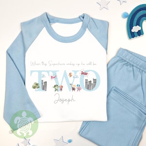May include: A light blue and white pajama set. The white top features the text "When this Superhero wakes up he will be TWO Joseph", with superhero graphics and cityscapes in the number "TWO".