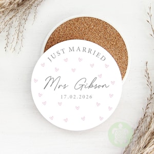 May include: A white round coaster with a cork base. The coaster has the text "JUST MARRIED" at the top, with the name "Mrs Gibson" and the date "17.02.2026" in the center. Small pink hearts are scattered around the text.