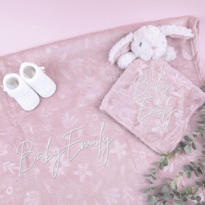 May include: A baby gift set featuring a pink floral blanket, a white plush bunny, a matching security blanket, and a pair of white baby booties. The name "Baby Emily" is embroidered on the blanket and security blanket.