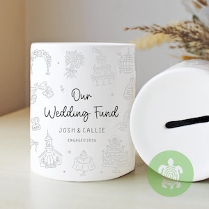 May include: White ceramic wedding fund bank with black text and illustrations. The text reads "Our Wedding Fund" with the names "Josh & Callie" and "Engaged 2026". The bank is decorated with wedding-themed line drawings.