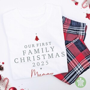 May include: A white t-shirt featuring the grey text "OUR FIRST FAMILY CHRISTMAS 2025" with a small red glittery Christmas tree icon above and the word "Mummy" in red script below, paired with red, black, and white plaid pajama pants.