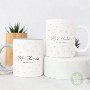 May include: Two white ceramic mugs with heart patterns and personalized text. One mug reads "Mr Thomas" and "08.09.2025", the other reads "Mrs Hudson" and "21.11.2025". The mugs have a simple, elegant design.