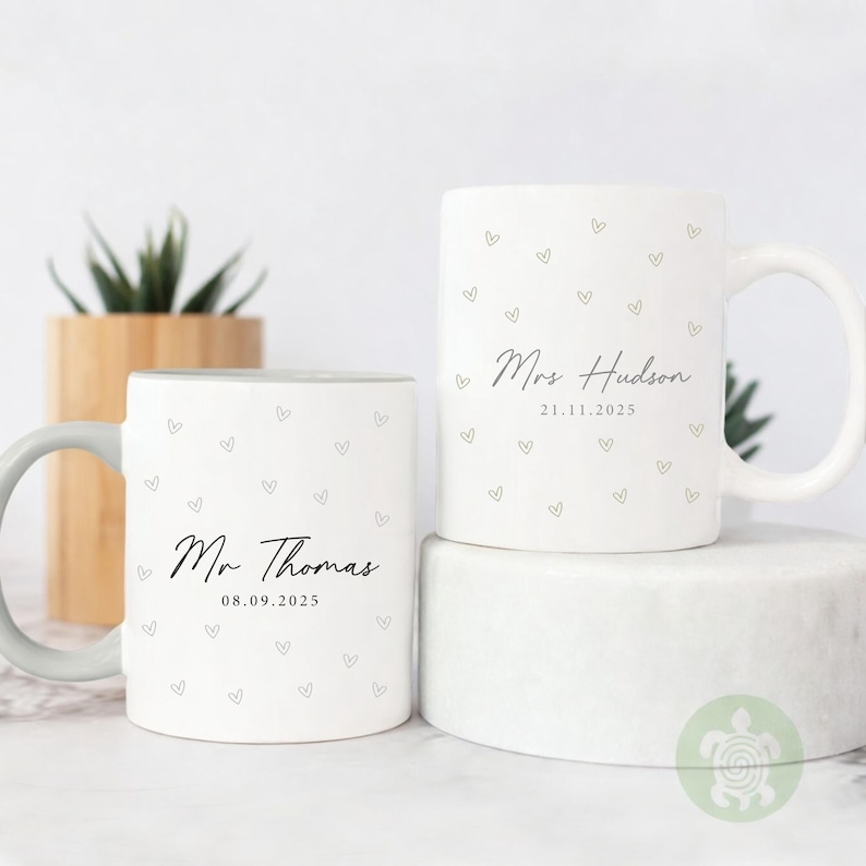 May include: Two white ceramic mugs with heart patterns and personalized text. One mug reads "Mr Thomas" and "08.09.2025", the other reads "Mrs Hudson" and "21.11.2025". The mugs have a simple, elegant design.