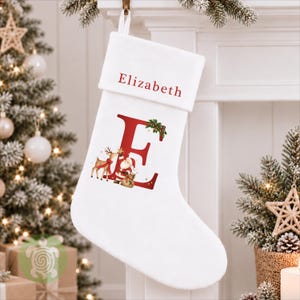 May include: A white Christmas stocking featuring a red embroidered name Elizabeth on the cuff. The centre displays a large red letter E decorated with a graphic of Santa Claus and a reindeer, accented with green holly leaves and red berries.