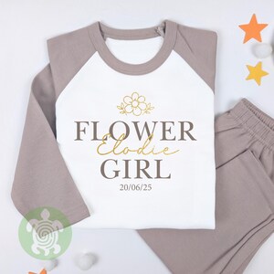 May include: A two-piece pyjama set with a white top and grey raglan sleeves, plus matching grey trousers. The top displays the words "FLOWER Elodie GIRL" in brown and gold script, a flower graphic, and the date 20/06/25.