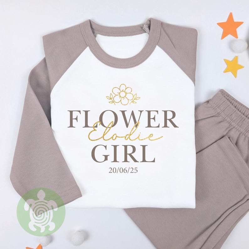 May include: A two-piece pyjama set with a white top and grey raglan sleeves, plus matching grey trousers. The top displays the words "FLOWER Elodie GIRL" in brown and gold script, a flower graphic, and the date 20/06/25.
