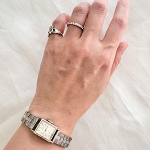 May include: A silver wristwatch with a square face and a silver metal band. The watch is on a person's wrist. The person is also wearing two silver rings on their fingers.