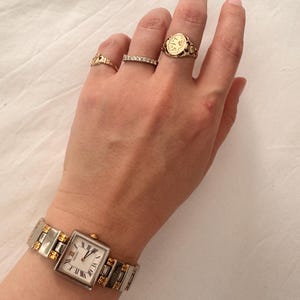 May include: A gold and silver wristwatch with a square face and Roman numerals. The watch is on a wrist with several gold rings, including a signet ring. The watch band is silver with gold accents.