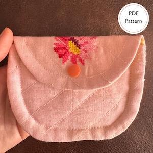 May include: A light pink quilted fabric pouch featuring a cross-stitched floral design in shades of pink, red, and yellow. The pouch has a rounded flap secured with a small orange circular snap button. A circular label reads PDF Pattern.