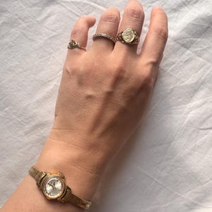 Può includere: Una collezione di gioielli color oro, tra cui anelli e un orologio da polso. Gli anelli presentano vari design, tra cui una corona, una fascia di piccole pietre e un anello con sigillo. L'orologio da polso ha un cinturino dorato e un quadrante argentato.